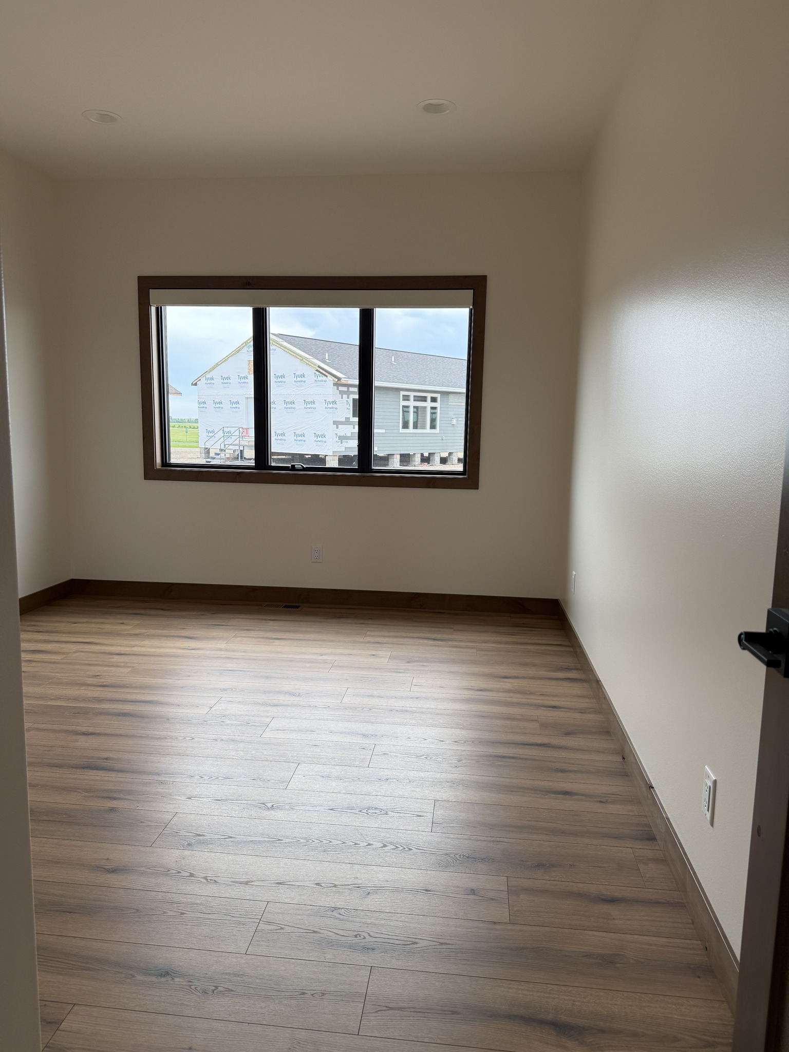 SEO-optimized alt text for a high-ranking modern luxury modular home bedroom featuring a professional interior design with warm cream walls and high-quality wide-plank oak flooring; the spacious room showcases a large black-framed window that provides a clear view of the residential construction site, complemented by elegant dark wood baseboards and contemporary black hardware on a solid wood door, perfectly highlighting premium prefab home floor plans, minimalist craftsman-style architectural details, and upscale durable residential mobile living finishes for maximum search engine visibility and ranking.