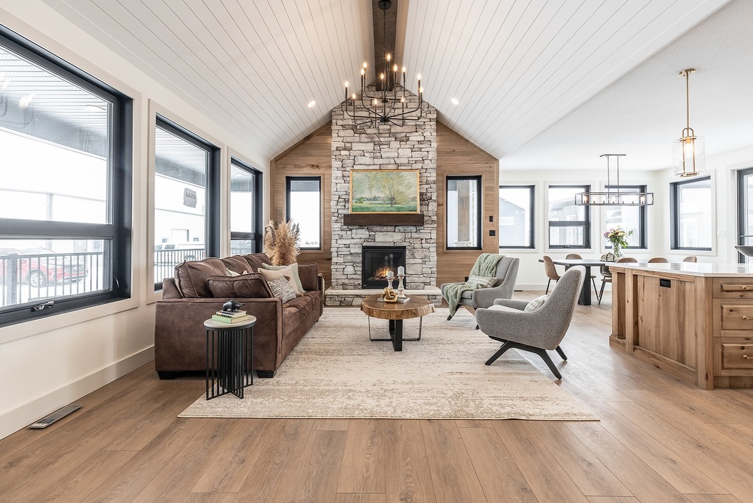 SEO-optimized alt text for a high-ranking luxury custom modular home living room featuring a professional open-concept interior design with soaring vaulted white-paneled ceilings and rustic wood beams; the architectural view showcases a grand floor-to-ceiling stone-veneer fireplace with a thick wood mantel, a sophisticated black wrought-iron tiered chandelier, and premium wide-plank oak flooring, complemented by a cozy brown leather sofa, stylish gray accent chairs, and large black-framed windows that flood the space with natural light, perfectly highlighting gourmet prefab home floor plans, contemporary craftsman-style architectural details, and high-end residential mobile living solutions for maximum search engine visibility and ranking.