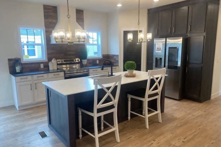 SEO-optimized alt text for a high-ranking luxury multi-section manufactured home kitchen featuring a professional gourmet interior design with a large central kitchen island and elegant white X-back barstools; the bright, open-concept floor plan showcases a sophisticated contrast between white shaker-style base cabinets and floor-to-ceiling dark espresso pantry cabinetry, a premium stainless steel French door refrigerator, and a modern range with a custom white vent hood set against a rustic wood-plank backsplash, all illuminated by contemporary gold-accented chandeliers and recessed lighting to highlight spacious double-wide mobile home floor plans, upscale modular kitchen decor, and luxury residential prefab home finishes for maximum search engine visibility and ranking.