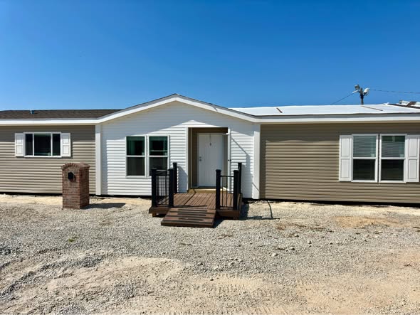 Exterior view of a modern double-wide manufactured home featuring tan horizontal vinyl siding with a contrasting white gabled entryway, white-trimmed windows with matching decorative shutters, and a small dark wood front porch with black railing, all set on a gravel foundation under a clear blue sky.