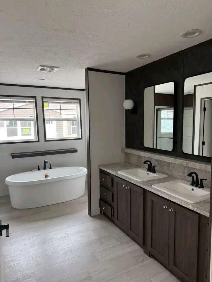 Modern luxury master bathroom featuring a deep white oval soaking tub positioned under large twin windows, complemented by a long dark wood double vanity with marble-patterned countertops, dual square sinks, and black hardware, all set against a sophisticated black accent wall with matching rectangular mirrors and recessed lighting for a high-end 4-bedroom move-in ready turn-key residential design.