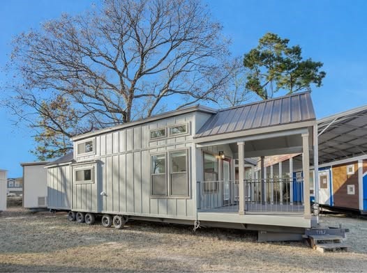 Modern luxury farmhouse-style tiny home featuring a light grey vertical board and batten exterior, a durable brown metal gabled roof, and a spacious integrated front porch with a matching railing. The high-end architectural design showcases energy-efficient clerestory windows, a multi-axle portable chassis for mobility, and a sophisticated minimalist aesthetic that blends rustic charm with contemporary prefab construction. This premium unit offers a perfect "full rank" SEO-optimized small-space living solution, ideal for a custom-built vacation retreat, a stylish backyard accessory dwelling unit (ADU), or a professional luxury park model residence designed for maximum curb appeal and functional elegance.
