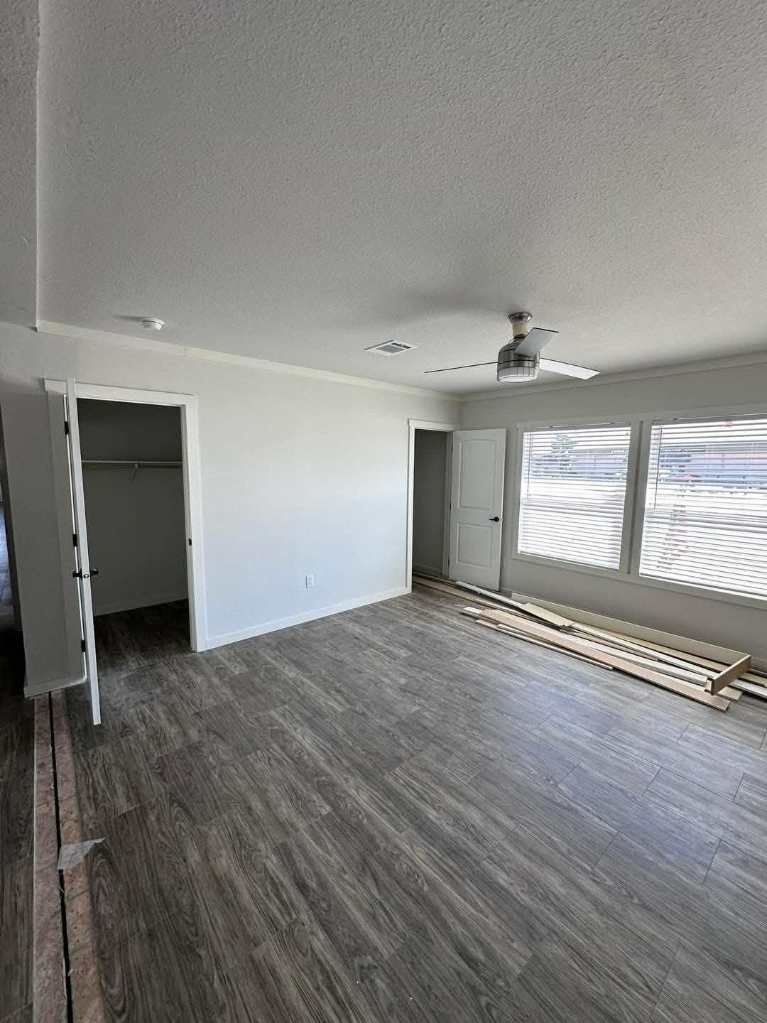Modern master bedroom interior in a new manufactured home featuring grey wood-look laminate flooring, neutral light grey walls, and large windows with white blinds, including a spacious walk-in closet and a contemporary brushed nickel ceiling fan for a clean, minimalist residential design.