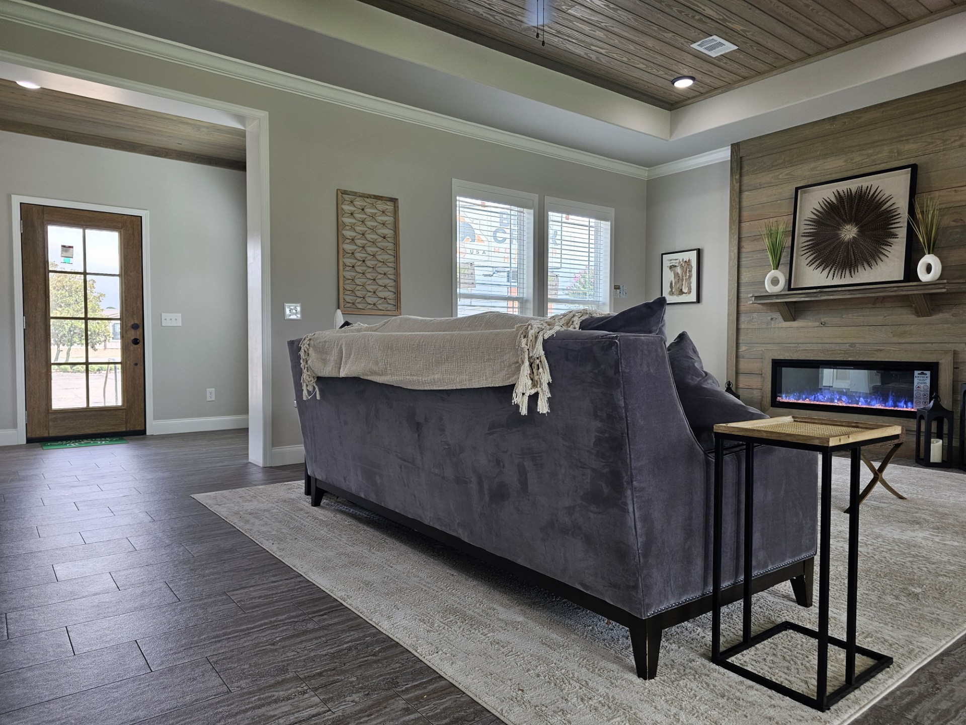 Modern luxury manufactured home interior featuring a spacious open-concept living room and gourmet kitchen with high-end tray ceilings finished in natural wood planks and a contemporary black ceiling fan. The sophisticated architectural design showcases a floor-to-ceiling reclaimed wood fireplace surround with an integrated linear electric fireplace, while the designer kitchen in the background features light grey cabinetry, a large center island with elegant pendant lighting, and a professional-style range hood. This "full rank" SEO-optimized layout highlights premium wood-look tile flooring, stylish transitional furniture, and an airy, upscale atmosphere, making it a perfect showcase for luxury prefab living, custom modular home floor plans, or modern permanent housing solutions designed with a high-end, contemporary farmhouse aesthetic.