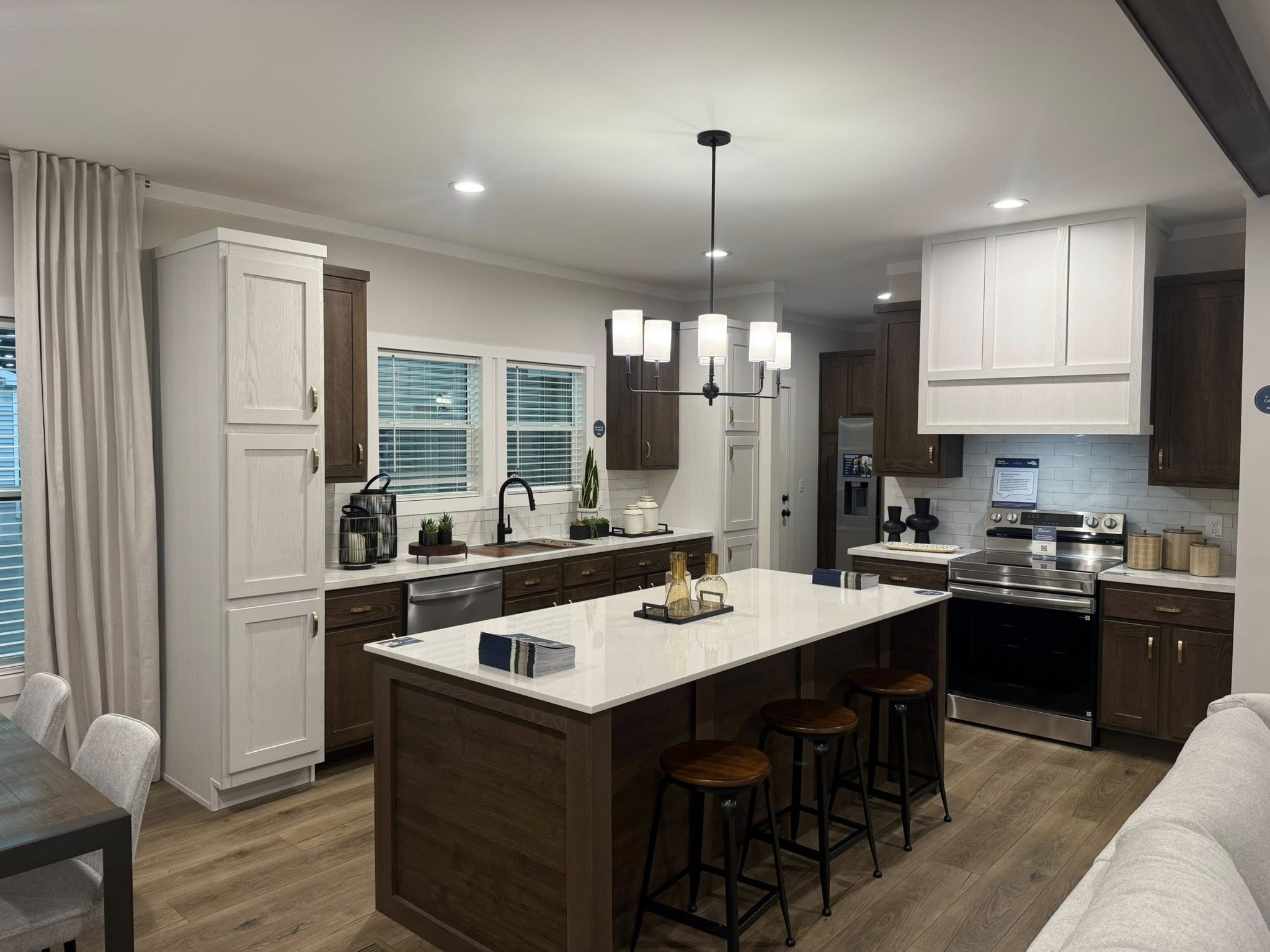 Modern kitchen with wooden floor, featuring a large island counter with white surface and wooden base, three barstools, and overhead lighting. Dark wood cabinets and stainless steel appliances enhance the sleek, contemporary look. Soft lighting creates a warm, inviting ambiance.