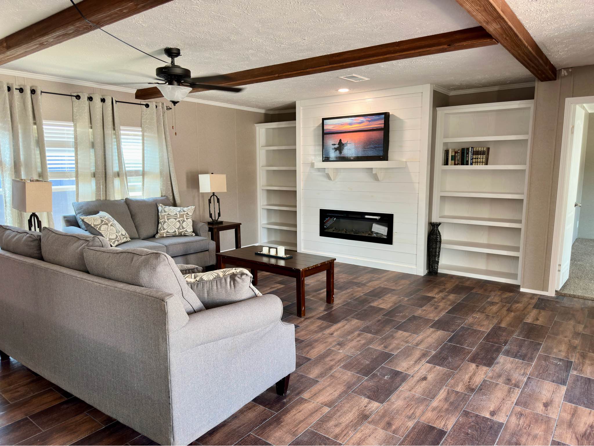 Modern farmhouse living room interior in a luxury manufactured home featuring a white shiplap fireplace wall with a built-in electric heater and wall-mounted TV, flanked by custom white built-in bookshelves, rustic dark wood ceiling beams, a black ceiling fan, grey upholstered sofas with patterned accent pillows, and dark wood-look plank flooring for a cozy, open-concept design.