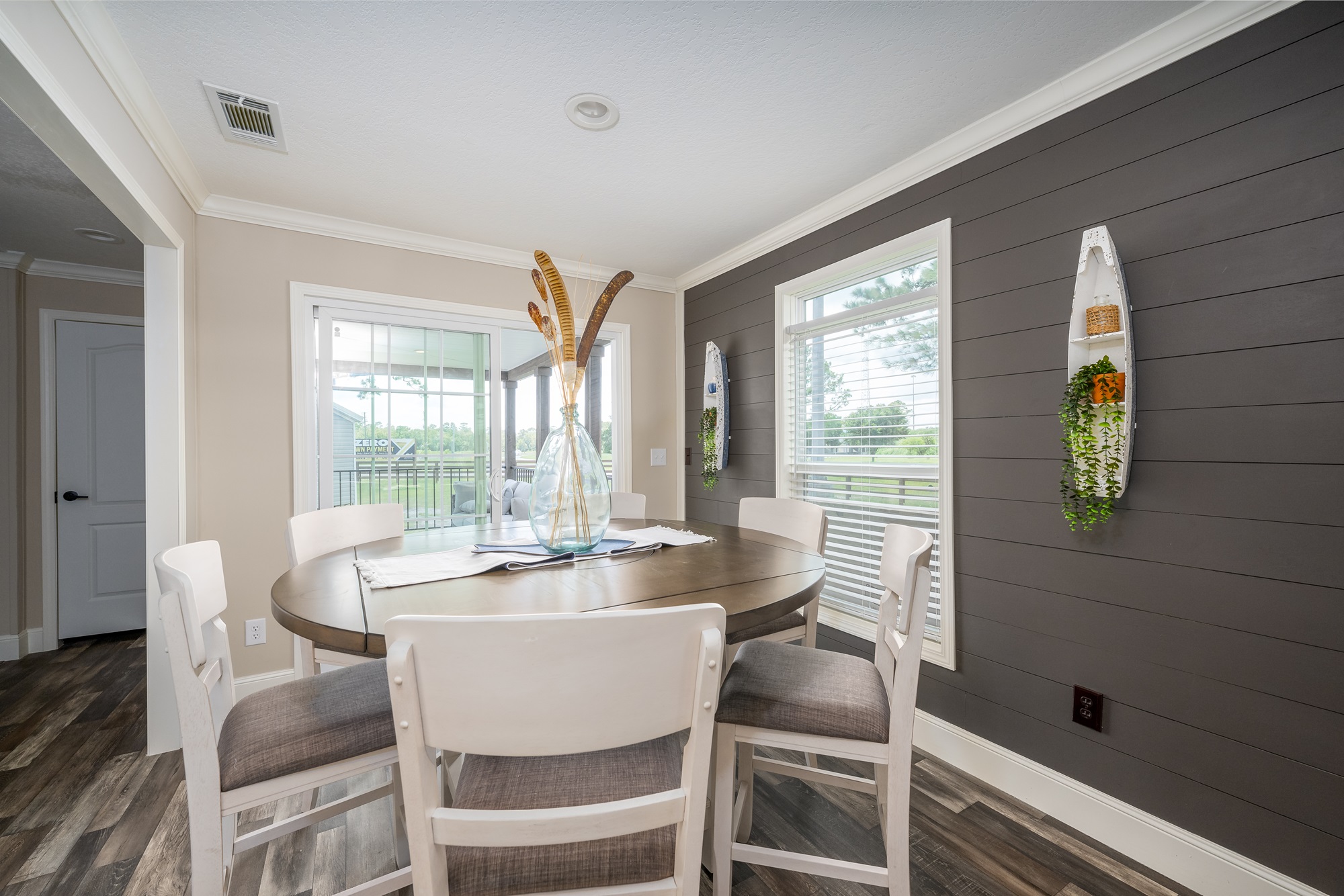 Cozy coastal farmhouse dining room in The Garnet 1,873 sq ft 3-bedroom customizable modular home featuring elegant round wood-grain dining table seating six with white upholstered chairs in neutral taupe fabric, dramatic dark gray shiplap accent wall with white trim, large picture windows draped in white blinds flooding the space with natural light and scenic outdoor views, centerpiece tall glass vase with dried pampas grass feathers, nautical wall decor including white wooden boat shelf with greenery and basket, recessed ceiling lights and ceiling fan, open doorway to porch and living areas, warm wood-look flooring creating an inviting bright family dining space perfect for meals and gatherings in this dream home giveaway winner.