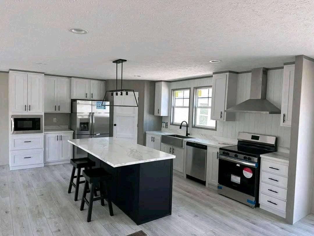 Modern spacious open-concept kitchen in luxury manufactured home featuring white shaker cabinets with black hardware, large central island with white marble-look quartz countertop and black base, black bar stools, stainless steel appliances including French door refrigerator microwave built-in oven dishwasher and electric range, farmhouse apron sink with matte black faucet, gray shiplap accent walls, white wood-look luxury vinyl plank flooring, black pendant lighting chandelier, recessed ceiling lights textured ceiling, bright natural light from large windows, contemporary farmhouse style modular home kitchen interior 2025 model