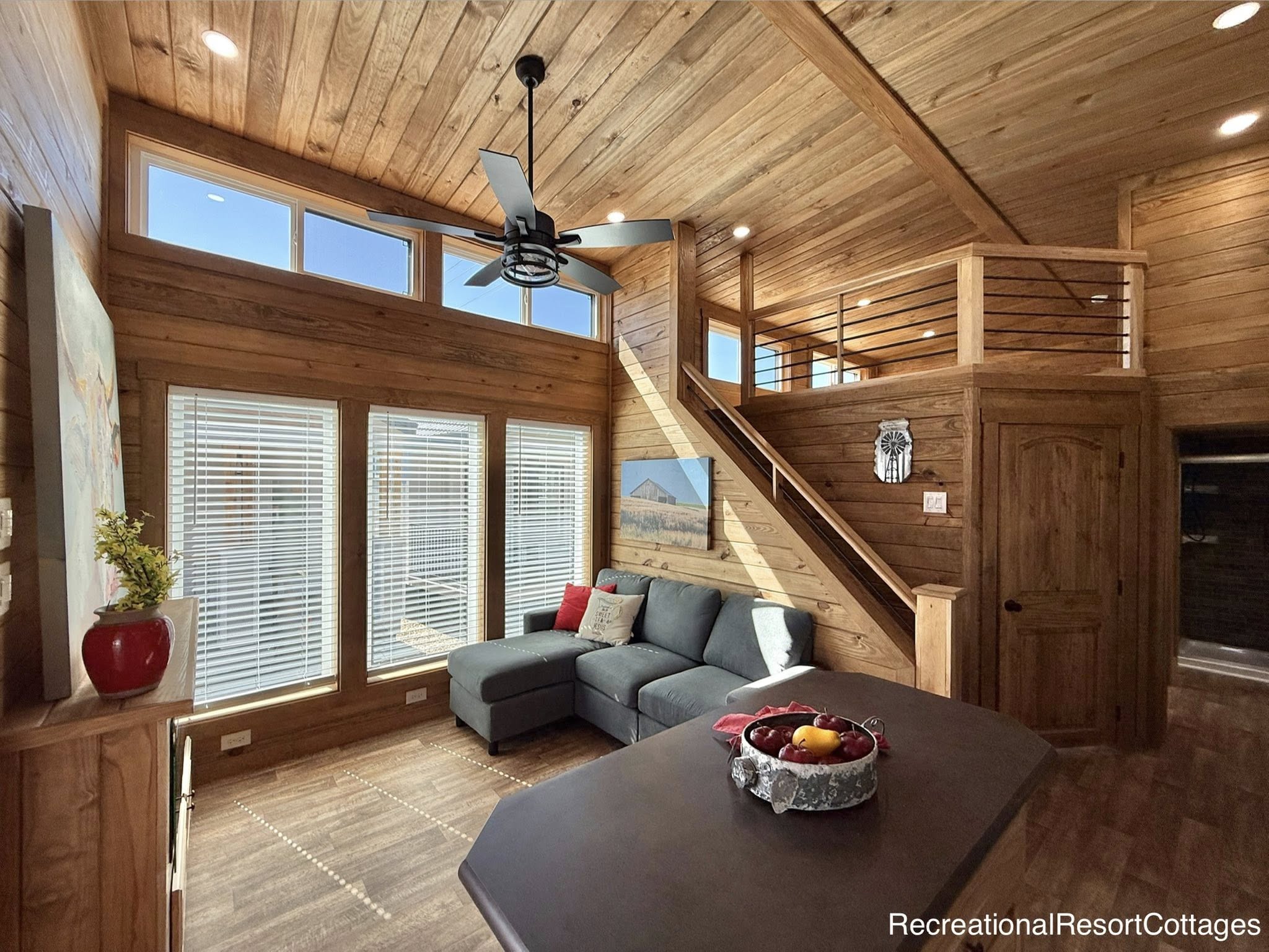 A cozy cabin interior with warm wood paneling, large windows, and a gray sectional sofa. A ceiling fan hangs above, and an open loft is visible.