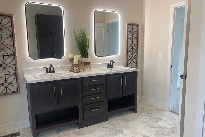 SEO-optimized alt text for a high-ranking luxury multi-section manufactured home master bathroom featuring a professional modern interior design with a large dark-wood double vanity; the sophisticated layout showcases premium shaker-style cabinetry with gold hardware, a crisp white countertop with dual integrated sinks and black faucets, and two contemporary backlit LED mirrors that provide a soft ambient glow, all perfectly complemented by high-end marble-patterned hexagonal floor tile to highlight spacious double-wide mobile home floor plans, upscale modular bathroom decor, and luxury residential prefab home finishes for maximum search engine visibility and ranking.