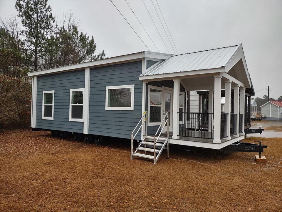 A small blue manufactured home with a white trim and metal roof sits on a brown grass lot. It has a covered porch with steps, under a cloudy sky.