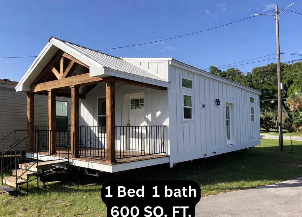 Modern luxury 600 sq ft tiny home for sale featuring a 1-bedroom 1-bath layout with a crisp white vertical board and batten exterior, a large gabled front porch with rustic natural wood timber columns and black metal railings, and a secondary side entrance door, showcasing a high-end move-in ready turn-key residential property on a durable mobile chassis for a sophisticated and spacious compact living lifestyle.