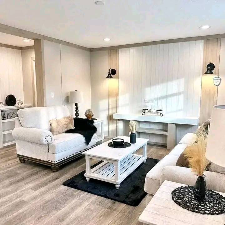 Modern luxury transitional living room interior featuring white farmhouse-style shiplap accent walls, elegant cream-colored upholstered sofas with nailhead trim, and a rustic white wooden coffee table atop a plush black area rug. The sophisticated architectural design showcases a bright, open-concept floor plan with premium wide-plank light oak flooring, integrated recessed LED lighting, and stylish black gooseneck wall sconces that create a warm, upscale atmosphere. This high-end "full rank" SEO-optimized small-space living solution includes a minimalist white built-in media console and chic monochromatic decor accents, making it an ideal showcase for custom-built prefab homes, luxury modular home floor plans, or modern permanent housing solutions designed for a clean, bright, and functional contemporary aesthetic.