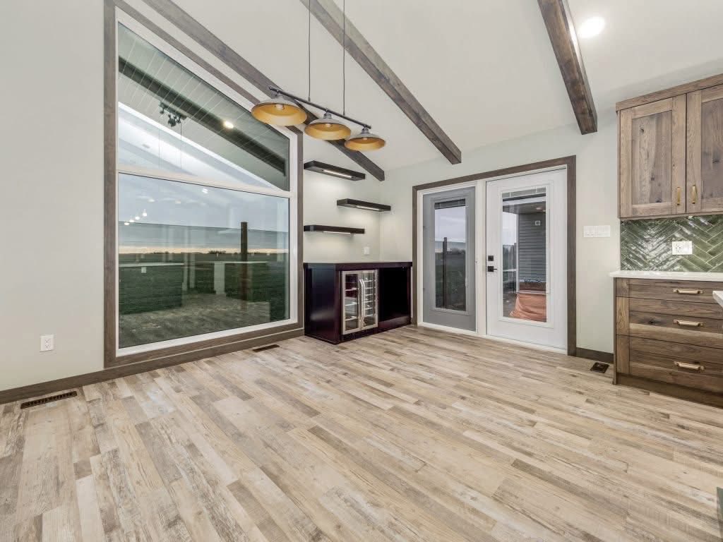 Modern open-concept living space with vaulted ceiling exposed wooden beams, large floor-to-ceiling panoramic windows overlooking scenic countryside view, luxury vinyl plank flooring in light oak finish, pendant track lighting with brass accents, built-in floating shelves, wine cooler station, French doors leading to outdoor deck, contemporary kitchen featuring shaker-style walnut cabinets, marble-look backsplash and quartz countertop – perfect luxury farmhouse interior design