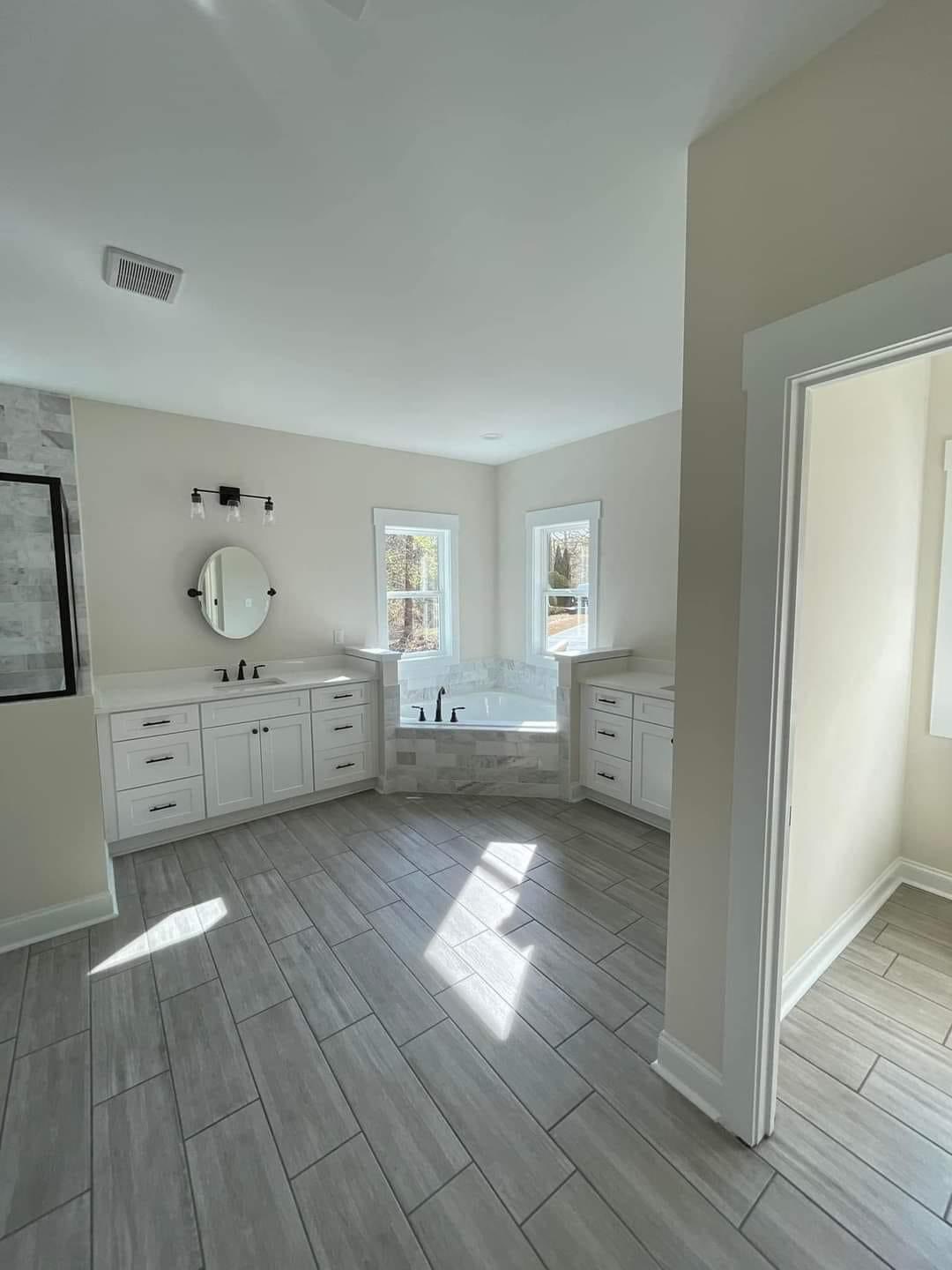 Spacious bathroom with light gray floor tiles, featuring a white double vanity, round mirror, and a corner bathtub beneath two sunlit windows.