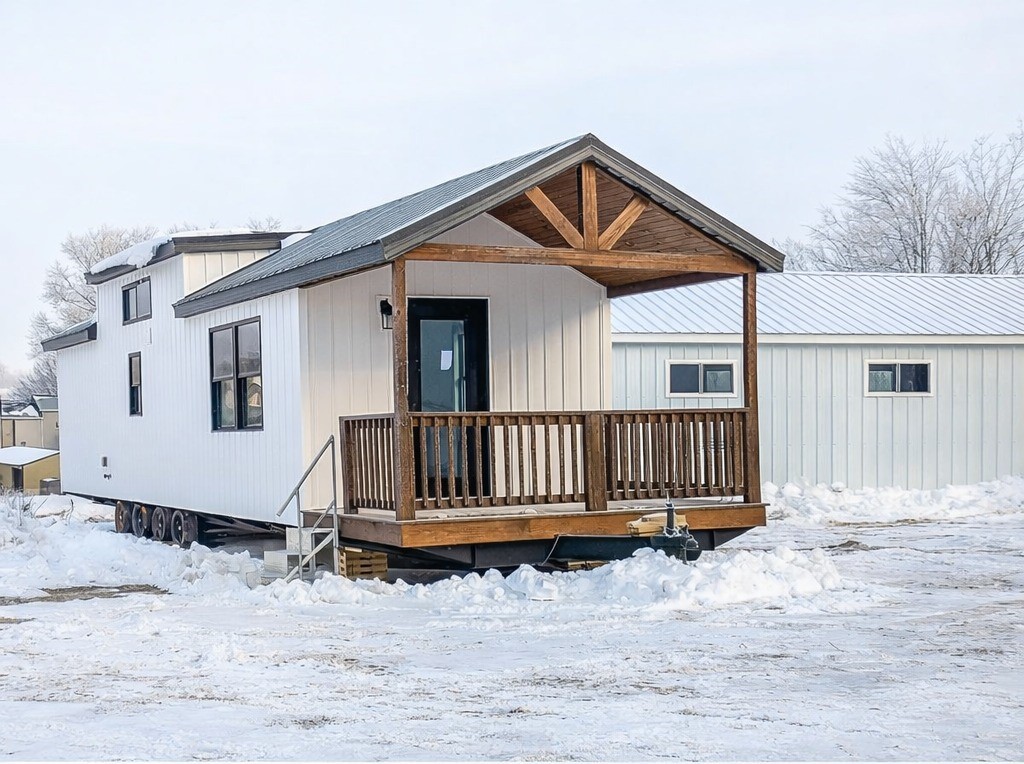 Modern luxury tiny home for sale in winter featuring a bright white vertical shiplap exterior with a large gabled front porch, rustic natural wood railings and timber beams, and a durable multi-axle mobile chassis, showcasing a high-end move-in ready turn-key residential design perfect for a sophisticated seasonal retreat or permanent compact living property.