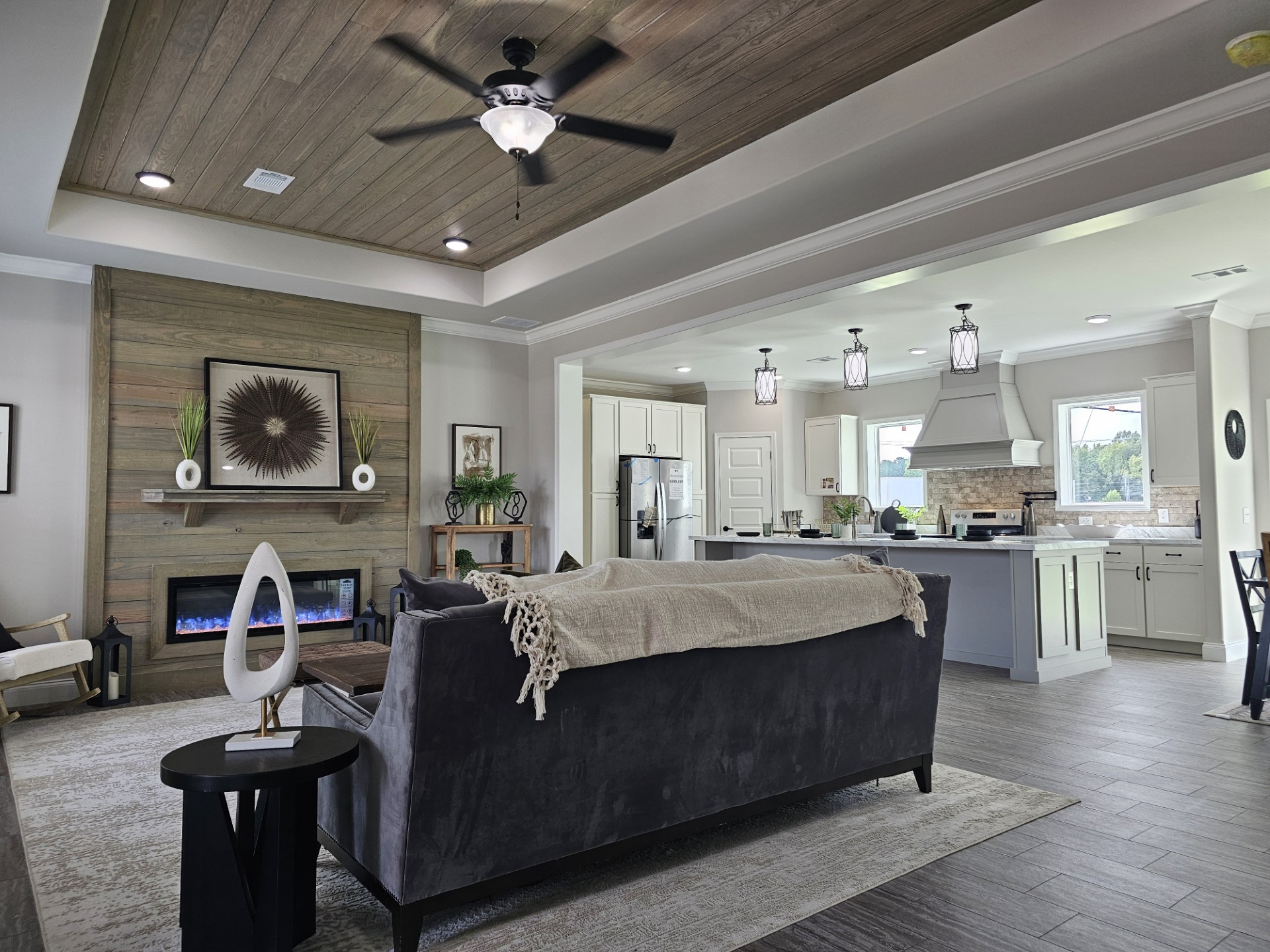 Modern luxury manufactured home interior featuring a spacious open-concept living room and gourmet kitchen with high-end tray ceilings finished in natural wood planks and a contemporary black ceiling fan. The sophisticated architectural design showcases a floor-to-ceiling reclaimed wood fireplace surround with an integrated linear electric fireplace, while the designer kitchen in the background features light grey cabinetry, a large center island with elegant pendant lighting, and a professional-style range hood. This "full rank" SEO-optimized layout highlights premium wood-look tile flooring, stylish transitional furniture, and an airy, upscale atmosphere, making it a perfect showcase for luxury prefab living, custom modular home floor plans, or modern permanent housing solutions designed with a high-end, contemporary farmhouse aesthetic.

Would you like me to create a list of focus keywords for this specific interior style?