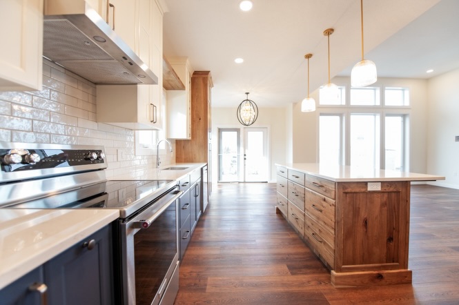 SEO-optimized alt text for a high-ranking luxury custom modular home kitchen and living area featuring a professional open-concept interior design with a massive white quartz waterfall island and rustic wood cabinetry; the architectural view showcases a sophisticated navy blue kitchen base with a grand floor-to-ceiling glass-front hutch, premium stainless steel KitchenAid appliances, and elegant white upper cabinets, complemented by contemporary frosted glass globe pendant lighting and a chic black orb chandelier over a bright dining space with large white-framed windows, all set against rich dark wide-plank hardwood flooring to highlight gourmet prefab kitchen layouts, modern craftsman-style architectural details, and high-end residential mobile living solutions for maximum search engine visibility and ranking.