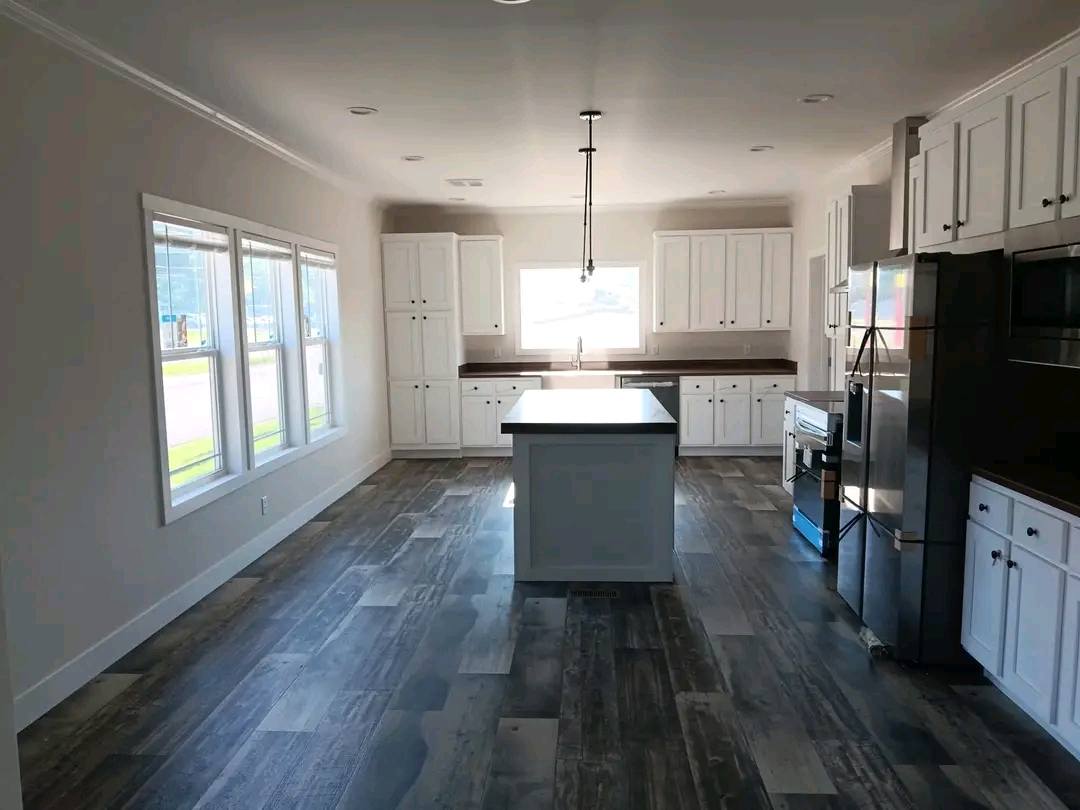 Modern luxury kitchen design featuring clean white shaker-style cabinetry with minimalist black hardware and a large central kitchen island with a dark countertop. The bright, open-concept interior showcases high-end stainless steel appliances, premium wide-plank wood-look flooring, and a large triple window that floods the space with natural light, perfectly capturing a sophisticated contemporary farmhouse aesthetic for a new modular or prefab home.