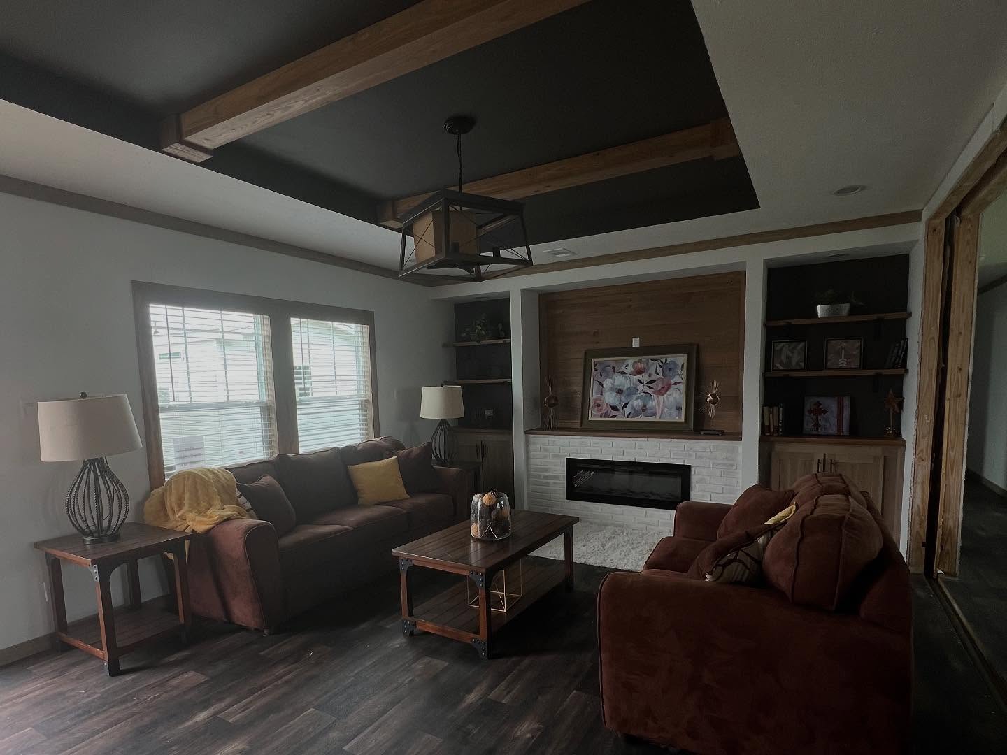 Cozy modern living room interior featuring plush chocolate brown sofas, a rustic wooden coffee table, and a white brick electric fireplace with wood-paneled accents and built-in shelving. The architectural design showcases a unique black tray ceiling with natural wood beams and a contemporary cage-style chandelier, complemented by large windows with blinds and dark wide-plank flooring for a sophisticated yet warm farmhouse aesthetic.