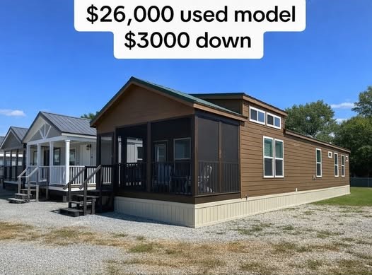 Modern luxury used park model home for sale featuring a brown horizontal lap siding exterior with a large screened-in front porch, black railings, and a tiered roofline with clerestory windows. This high-end prefab house is displayed on a gravel lot alongside other contemporary mobile units, highlighting a spacious and affordable small-space living solution for $26,000, perfect for full-rank SEO-optimized searches for custom vacation cottages, backyard accessory dwelling units (ADUs), or premium manufactured homes with significant curb appeal.