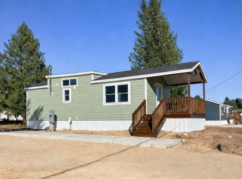 Modern luxury park model tiny home featuring a sophisticated sage green horizontal lap siding exterior, a dark gabled roof, and a prominent covered front porch with rich natural wood railings and a matching staircase. The contemporary architectural design includes a white corrugated metal skirting base, energy-efficient white-trimmed windows, and a spacious wood-plank deck, offering a premium and stylish small-space living solution perfect for a high-end prefab vacation cottage or custom-built accessory dwelling unit with exceptional curb appeal.