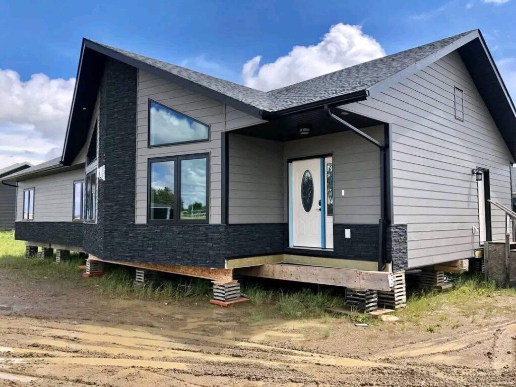 Gemini said Modern luxury contemporary manufactured home exterior featuring a sophisticated two-tone design with light grey horizontal siding and premium black stacked stone accents on a professional pier-and-beam foundation. The high-end architectural design showcases a unique asymmetrical gabled roofline with black trim, energy-efficient geometric windows that include a large triangular transom for maximum natural light, and a stylish white entry door with a decorative glass insert. This "full rank" SEO-optimized residence highlights exceptional curb appeal and high-quality craftsmanship, making it an ideal showcase for luxury prefab living, custom-built modular home floor plans, or modern permanent housing solutions designed with a bold, upscale, and contemporary transitional aesthetic.