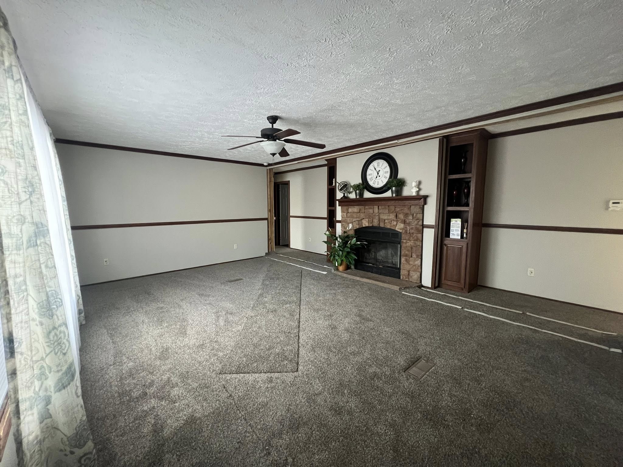 SEO-optimized alt text for a spacious, traditional manufactured home living room featuring neutral gray carpeting and a classic stone-surround fireplace with a rustic wood mantel; the high-ranking interior design showcases dark wood built-in bookshelves, a decorative wall clock, and a central ceiling fan with dark blades, all framed by elegant crown molding and floral-patterned curtains that highlight comfortable family-style modular home floor plans and affordable luxury mobile home decor for maximum search engine visibility.