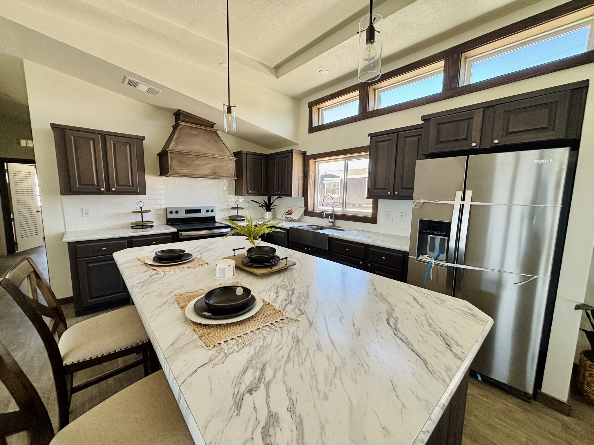 Modern kitchen with a large marble island, black cabinets, stainless steel appliances, and high windows. Plates set neatly, creating a welcoming vibe.