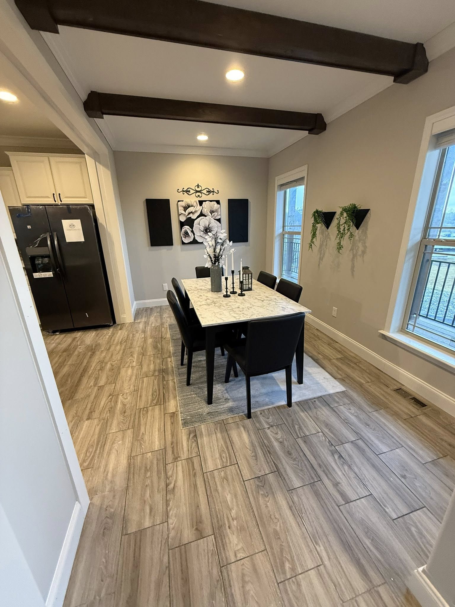 Luxury modern farmhouse dining room in premium tiny home or modular prefab cottage featuring vaulted tray ceiling with dramatic exposed dark wooden beams, recessed LED lighting, spacious open layout with seamless flow to adjacent kitchen, elegant marble-look quartz dining table centerpiece with black candlestick holders, gray vase floral arrangement, six black upholstered dining chairs, light gray accent walls adorned with black and white botanical canvas art and wrought iron plant shelves with greenery, wide windows with white trim flooding room with natural light and scenic views, warm gray-brown luxury vinyl plank flooring, sophisticated neutral palette creating inviting family gathering space perfect for contemporary ranch-style manufactured home, vacation cottage, or rural retreat interior design