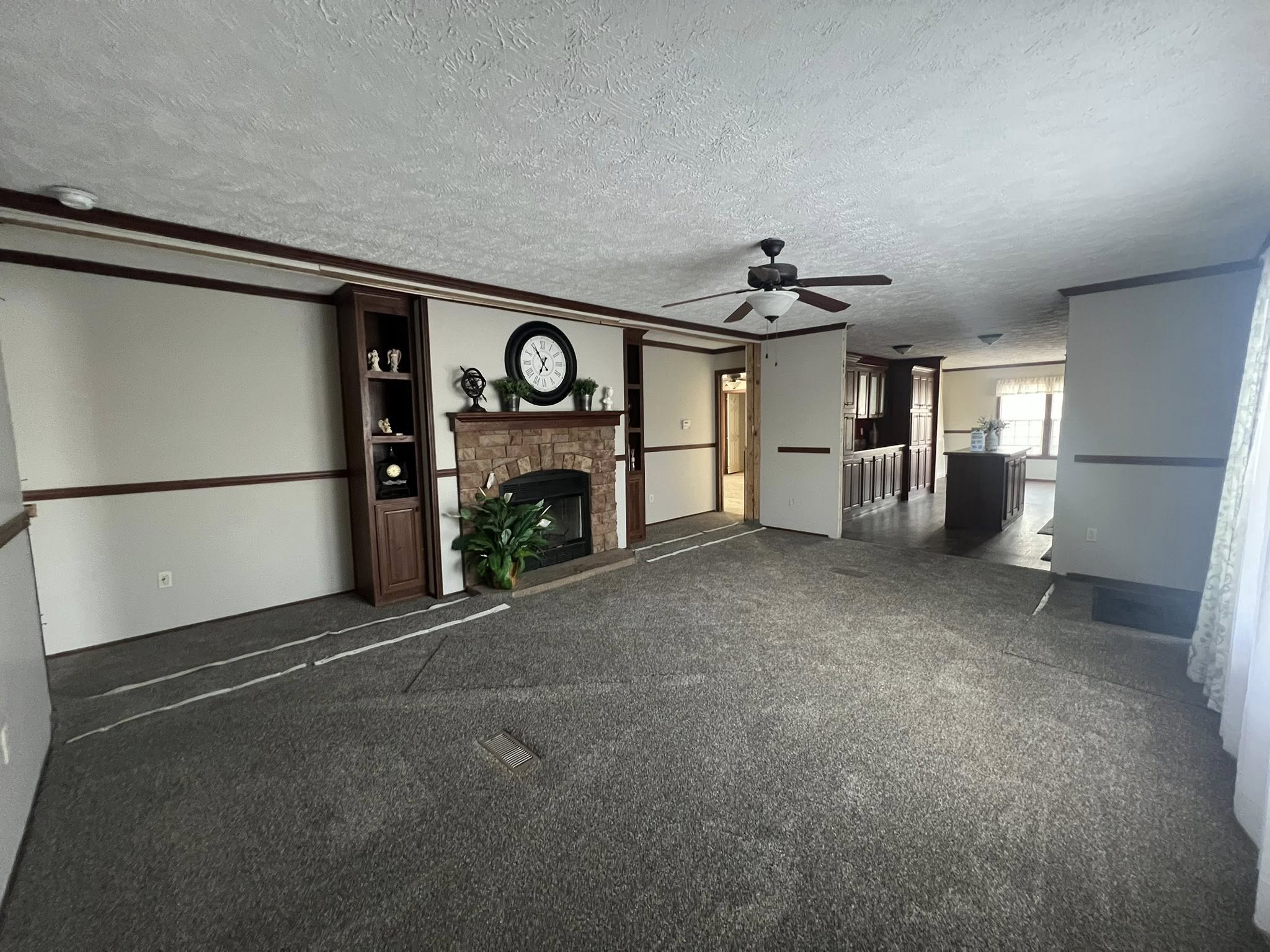 SEO-optimized alt text for a spacious, traditional manufactured home living room featuring neutral gray carpeting and a classic stone-surround fireplace with a rustic wood mantel; the high-ranking interior design showcases dark wood built-in bookshelves, a decorative wall clock, and a central ceiling fan with dark blades, all framed by elegant crown molding and floral-patterned curtains that highlight comfortable family-style modular home floor plans and affordable luxury mobile home decor for maximum search engine visibility.