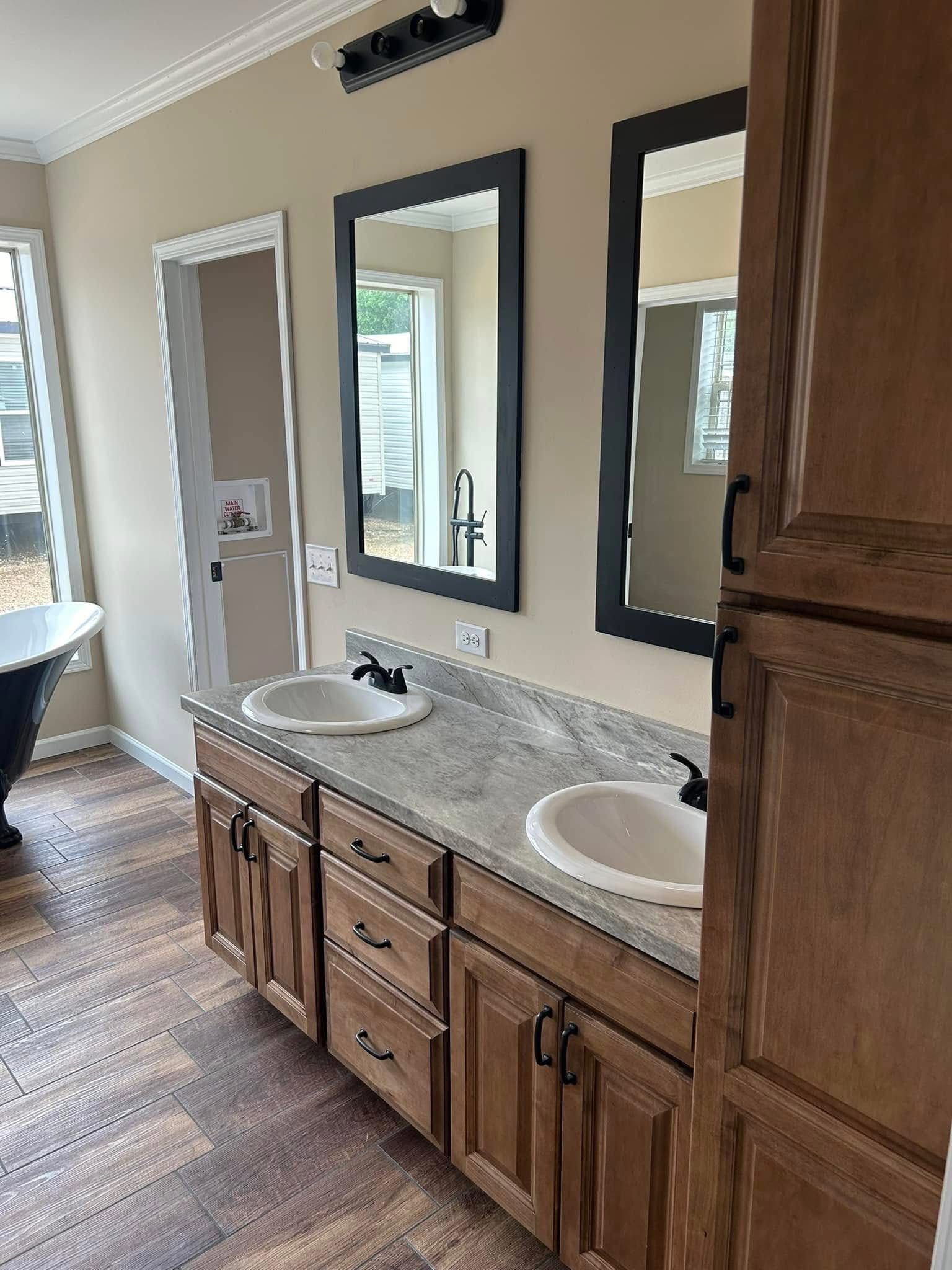 Luxurious bathroom featuring a freestanding clawfoot bathtub, a dual vanity with a granite-look countertop, two white undermount sinks with black faucets, framed mirrors, and a tall wooden cabinet, all set against light beige walls and dark wood-look tile flooring.