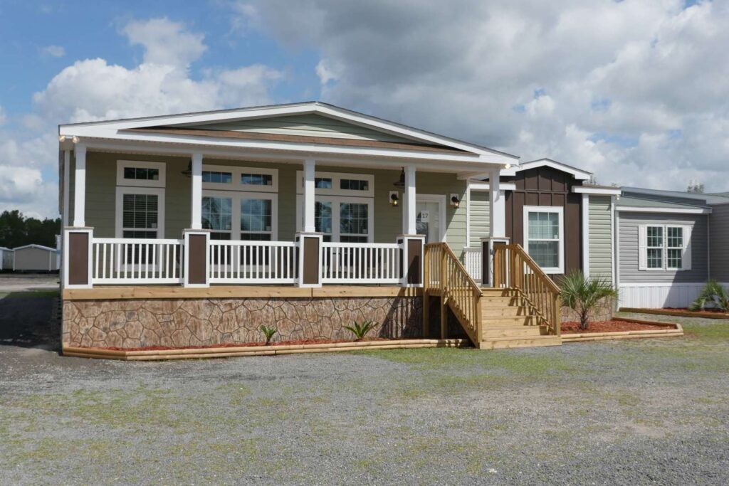 Modern manufactured home with a welcoming covered porch, featuring light green siding, white railings, dark brown accents, a stone-patterned foundation, and a sturdy wooden staircase leading to the entrance, set against a cloudy sky.