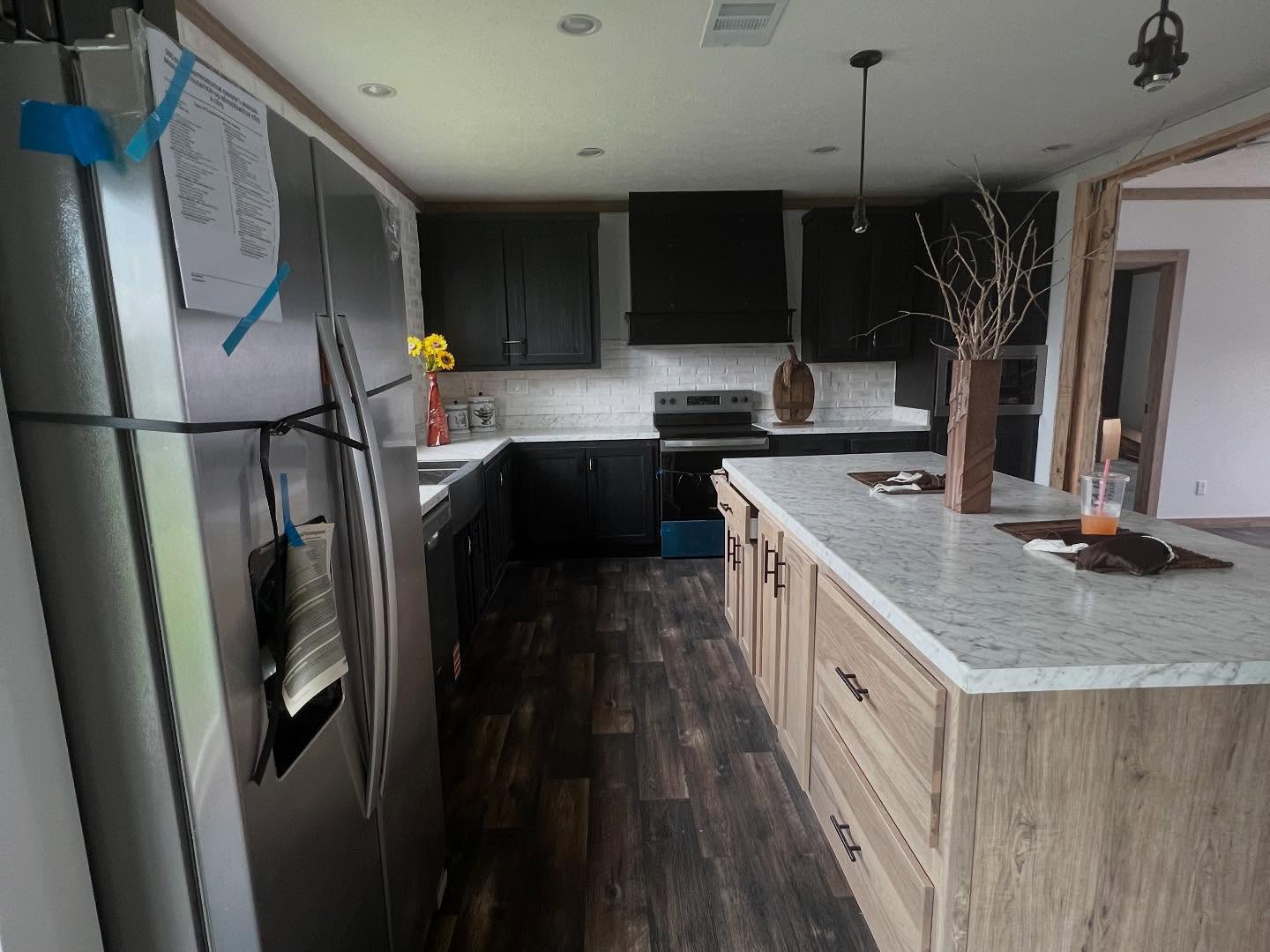Modern farmhouse kitchen design featuring dark charcoal upper cabinets and a black range hood paired with a large light wood island topped with white marble countertops. The open-concept space showcases a contemporary rustic aesthetic with dark wood flooring, a white subway tile backsplash, and stainless steel appliances, providing a luxury look for a modern modular or prefab home interior.