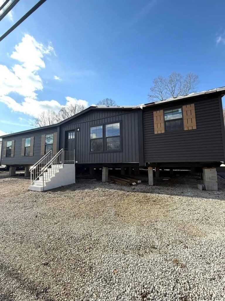 Modern luxury double-wide manufactured home for sale featuring a charcoal grey vertical board and batten siding exterior with a large front-facing gabled section, contrasting brown wood-style shutters, and a white concrete entry staircase on a professional block foundation for a sophisticated and affordable move-in ready turn-key residential property.