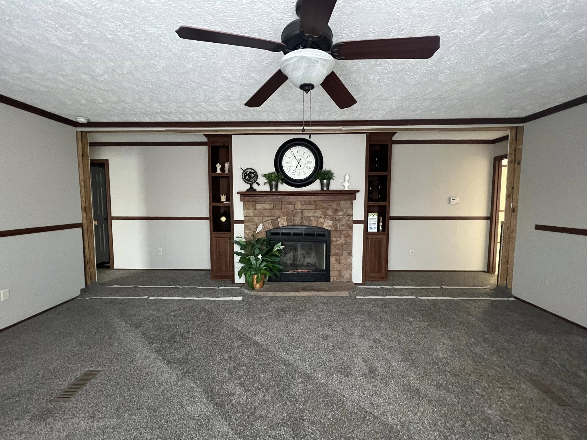 SEO-optimized alt text for a spacious, traditional manufactured home living room featuring neutral gray carpeting and a classic stone-surround fireplace with a rustic wood mantel; the high-ranking interior design showcases dark wood built-in bookshelves, a decorative wall clock, and a central ceiling fan with dark blades, all framed by elegant crown molding and floral-patterned curtains that highlight comfortable family-style modular home floor plans and affordable luxury mobile home decor for maximum search engine visibility.