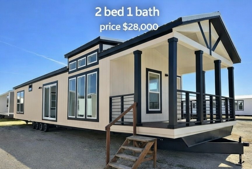Affordable modern tiny home exterior featuring a charming 2 bedroom 1 bath manufactured modular design priced at $28,000 with crisp beige lap siding contrasting bold black trim accents gabled roofline black-framed dormer windows spacious wraparound covered porch supported by sturdy black columns black metal railings wooden deck stairs leading to entry natural light streaming through large energy-efficient windows elevated foundation on trailer chassis under clear blue sky ideal compact family starter home cabin retreat or downsizing solution with massive curb appeal and outdoor living space in this budget-friendly dream tiny house model.