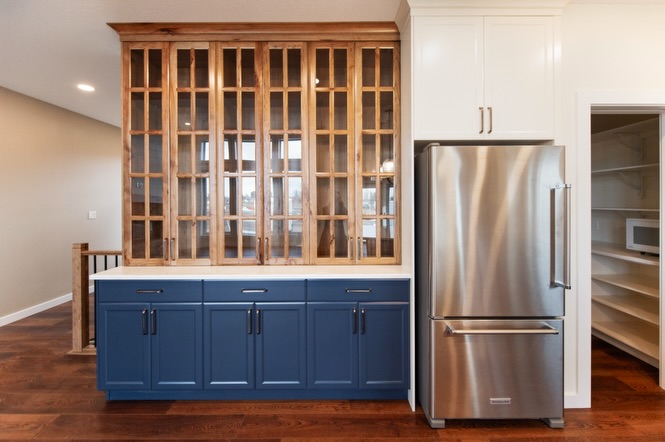 SEO-optimized alt text for a high-ranking luxury custom modular home kitchen featuring a professional open-concept interior design with a striking navy blue and rustic wood cabinetry combination; the architectural view showcases a grand floor-to-ceiling wood-framed glass-front hutch and premium stainless steel KitchenAid appliances, all complemented by elegant white shaker upper cabinets, a classic white subway tile backsplash, and contemporary gold pendant lighting set against rich dark wide-plank hardwood flooring, perfectly highlighting gourmet prefab kitchen layouts, modern craftsman-style architectural details, and high-end residential mobile living solutions for maximum search engine visibility and ranking.