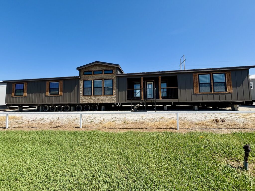 A large, dark-colored manufactured home with rustic wooden shutters sits on a gravel lot under a clear blue sky, with green grass in the foreground.