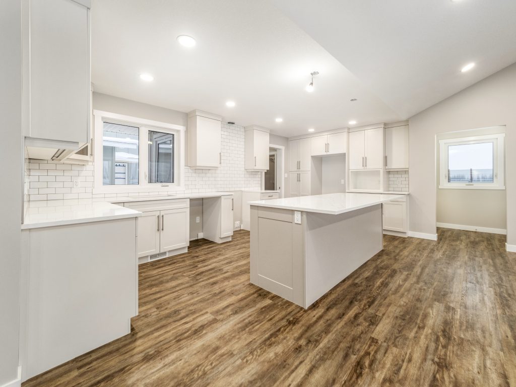 Luxurious spacious open-concept kitchen interior in premium modern manufactured home, bright white shaker cabinets with brushed nickel hardware throughout, massive center island with white quartz waterfall countertop and extended breakfast bar, white subway tile backsplash, stainless steel range hood over cooktop, built-in appliances including dishwasher and microwave, dark wood-look luxury vinyl plank flooring, recessed LED ceiling lights and modern pendant fixtures, large windows and sliding doors flooding natural light, airy high-end unfurnished gourmet kitchen layout in 2025 double-wide modular home design