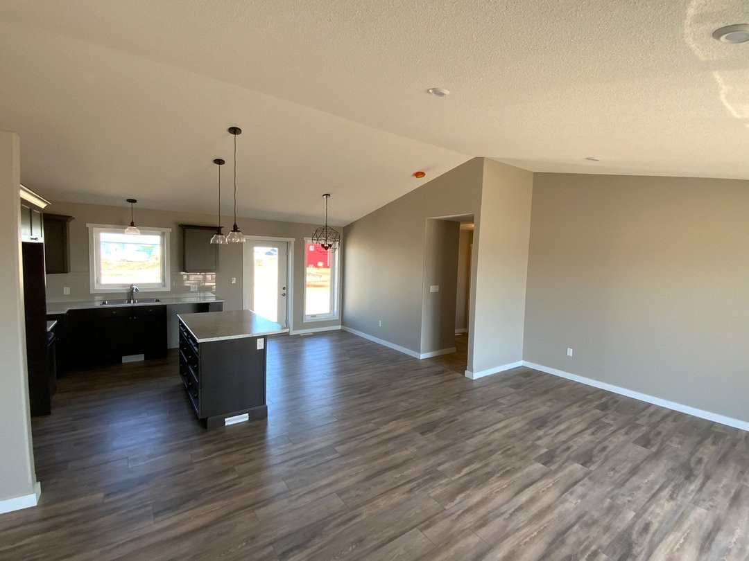 Modern luxury modular home interior featuring a spacious open-concept living and dining area with premium wide-plank wood flooring and neutral warm-toned walls, showcasing a contemporary kitchen with dark cabinetry, a functional center island, and stylish pendant lighting under a bright vaulted ceiling for a high-end move-in ready turn-key residential property design.