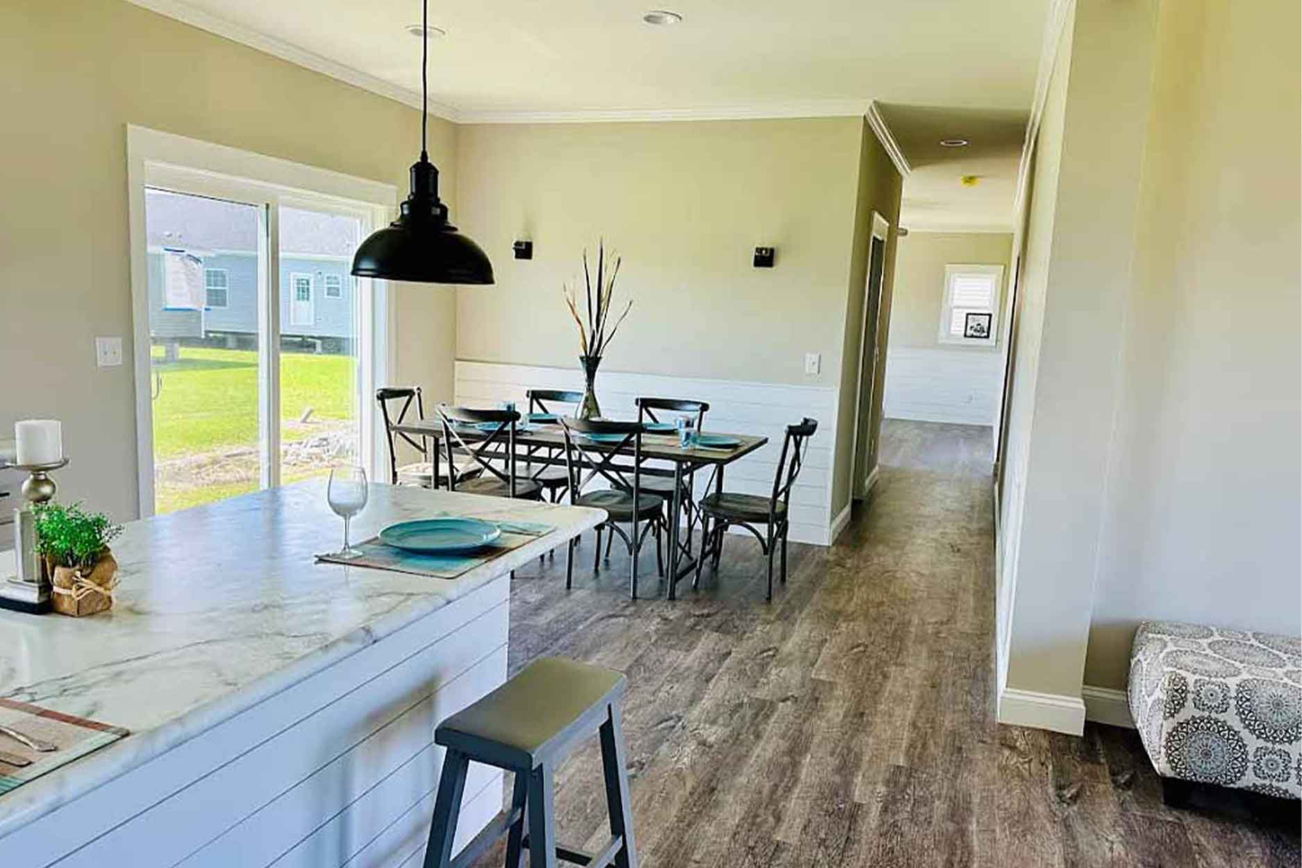 Modern luxury open-concept kitchen and dining area featuring a large white marble-topped island with grey barstools and black industrial pendant lighting. The bright interior showcases elegant white shaker-style cabinetry with black hardware, high-end stainless steel appliances, and a grey wide-plank wood floor that leads into a sunlit dining space with a dark wood table and cross-back chairs, perfectly capturing a sophisticated contemporary farmhouse aesthetic for a modular or prefab home.