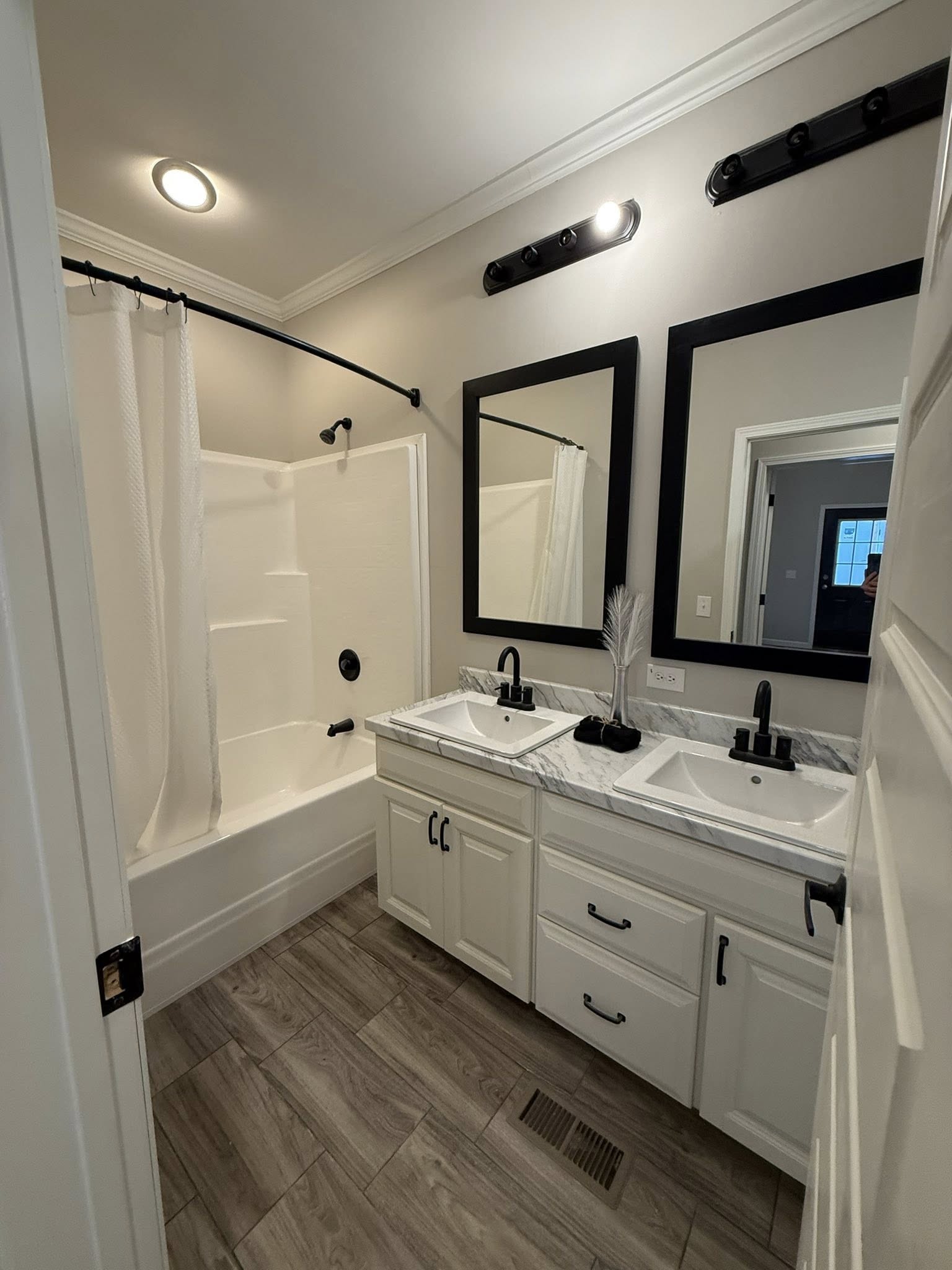 Luxury modern farmhouse bathroom in premium tiny home or modular prefab cottage featuring elegant double vanity with white shaker cabinets, marble-look quartz countertop, matte black widespread faucets and hardware, dual rectangular black-framed mirrors with industrial black barn door-style vanity light bar featuring exposed bulbs, spacious walk-in shower with white subway tile surround and curved shower rod with white curtain, deep soaking bathtub combo, light gray wood-look luxury vinyl plank flooring, creamy white walls with crown molding and tray ceiling accents, recessed lighting creating spa-like ambiance, pampas grass decor in clear vase, timeless neutral palette perfect for contemporary ranch-style manufactured home, vacation retreat, or family ensuite design