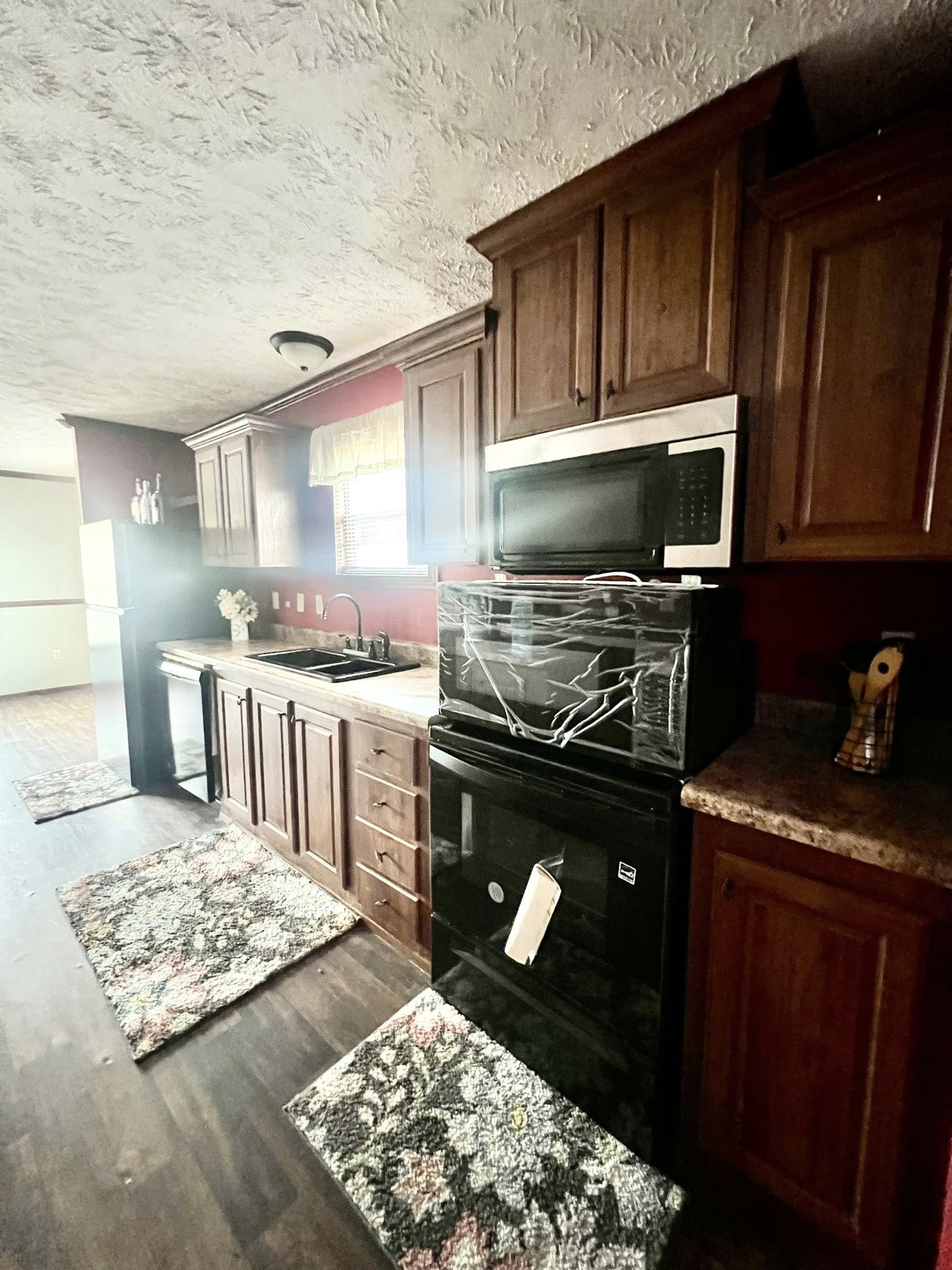 SEO-optimized alt text for a spacious manufactured home kitchen featuring extensive dark wood cabinetry, a central kitchen island, and a galley-style layout with modern black appliances; the high-ranking interior design showcases a functional and elegant floor plan with durable wood-look flooring, red accent walls, and recessed lighting that opens into a bright dining area, perfectly highlighting premium mobile home kitchen renovations and sophisticated modular home decor for maximum search engine visibility and ranking.