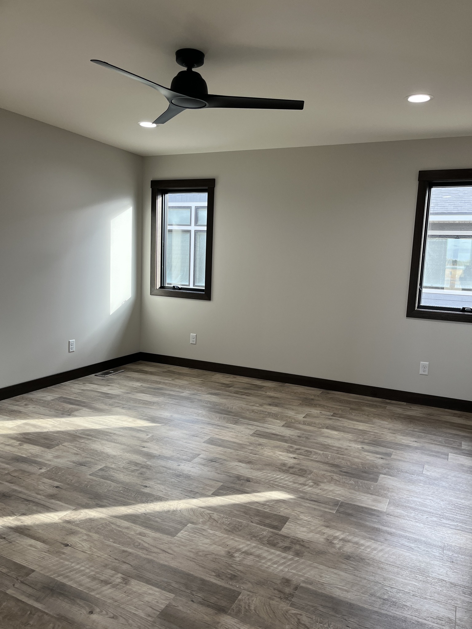 Modern luxury contemporary manufactured home interior featuring a spacious, empty room with premium wide-plank grey wood laminate flooring and minimalist light grey walls. The sophisticated architectural design includes black-trimmed energy-efficient windows that flood the space with natural light, a sleek black three-blade ceiling fan, and integrated recessed LED lighting for a clean, professional finish. This "full rank" SEO-optimized space highlights high-quality craftsmanship and modern aesthetics, making it an ideal showcase for luxury prefab living, custom modular home floor plans, or stylish permanent housing solutions designed with a minimalist and upscale transitional feel.