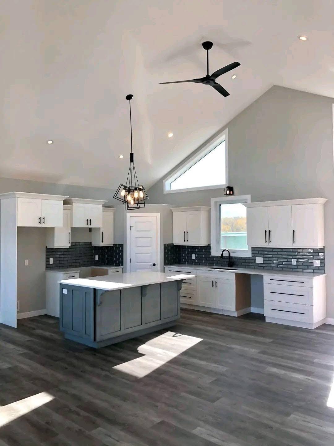 Modern luxury contemporary manufactured home kitchen featuring a large slate grey center island with a white quartz countertop, set against a backdrop of crisp white shaker cabinetry and a sophisticated dark grey subway tile backsplash. The high-end architectural design showcases a vaulted ceiling with a minimalist black three-blade ceiling fan, industrial-style black geometric pendant lighting, and a unique triangular transom window that maximizes natural light. This professional "full rank" SEO-optimized space includes premium wide-plank grey wood flooring, sleek black hardware, and integrated recessed LED lighting, making it an ideal showcase for luxury prefab living, custom modular home floor plans, or modern permanent housing solutions designed with a bright, upscale, and transitional aesthetic.