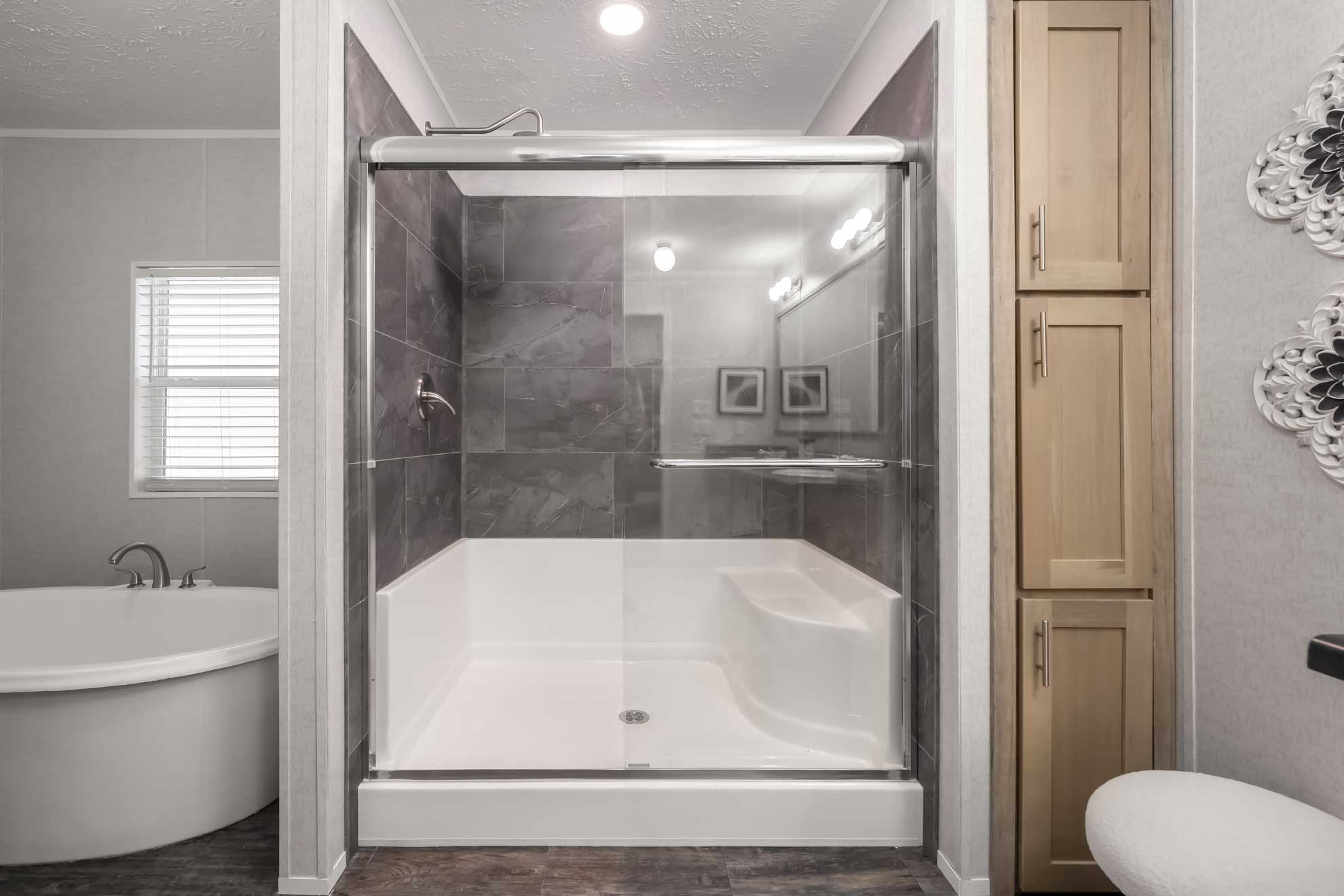 SEO-optimized alt text for a modern master bathroom in a luxury manufactured home featuring a large walk-in shower with charcoal gray tile and sliding glass doors, a deep soaking bathtub, and a light wood-finish linen cabinet; the high-ranking interior design showcases clean lines with neutral walls and durable wood-look flooring, perfectly highlighting premium mobile home bathroom renovations and contemporary modular home floor plans for maximum search engine visibility and ranking.