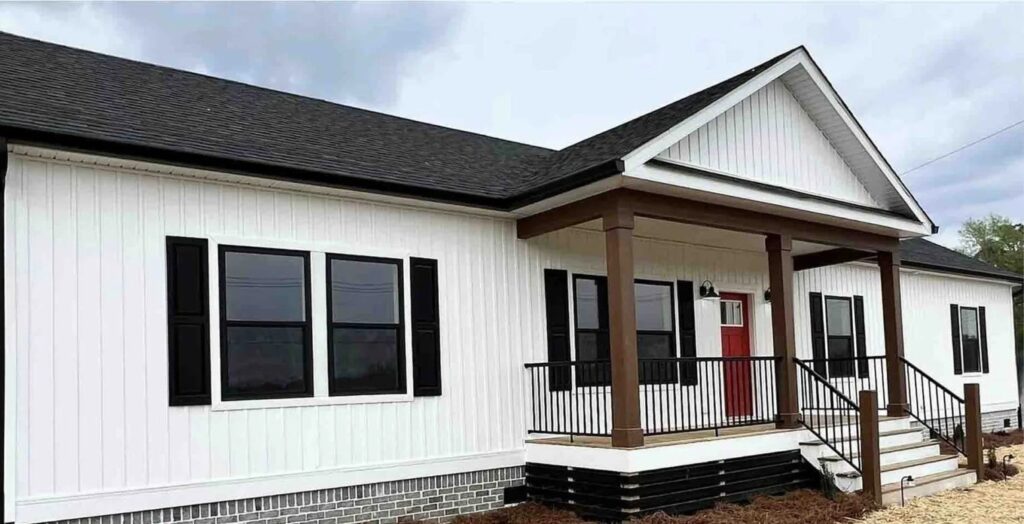 Modern white farmhouse-style modular home featuring white vertical board and batten siding, a black shingle roof, and a striking red front door. The exterior architectural design includes a spacious covered front porch with dark wood pillars, black metal railings, and black shutters on large windows, all set against a grey brick foundation for a high-end contemporary curb appeal.