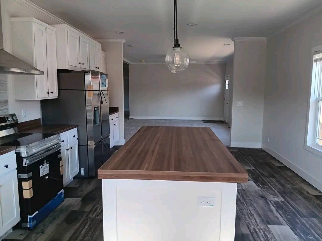 Modern luxury kitchen featuring elegant white shaker cabinets, a large central island with a butcher block countertop, and high-end stainless steel appliances including a double-door refrigerator. The contemporary open-concept interior design is highlighted by premium wide-plank grey wood flooring, minimalist pendant lighting, and bright neutral walls, creating a spacious and functional heart of the home perfect for modern modular or custom-built residential living.