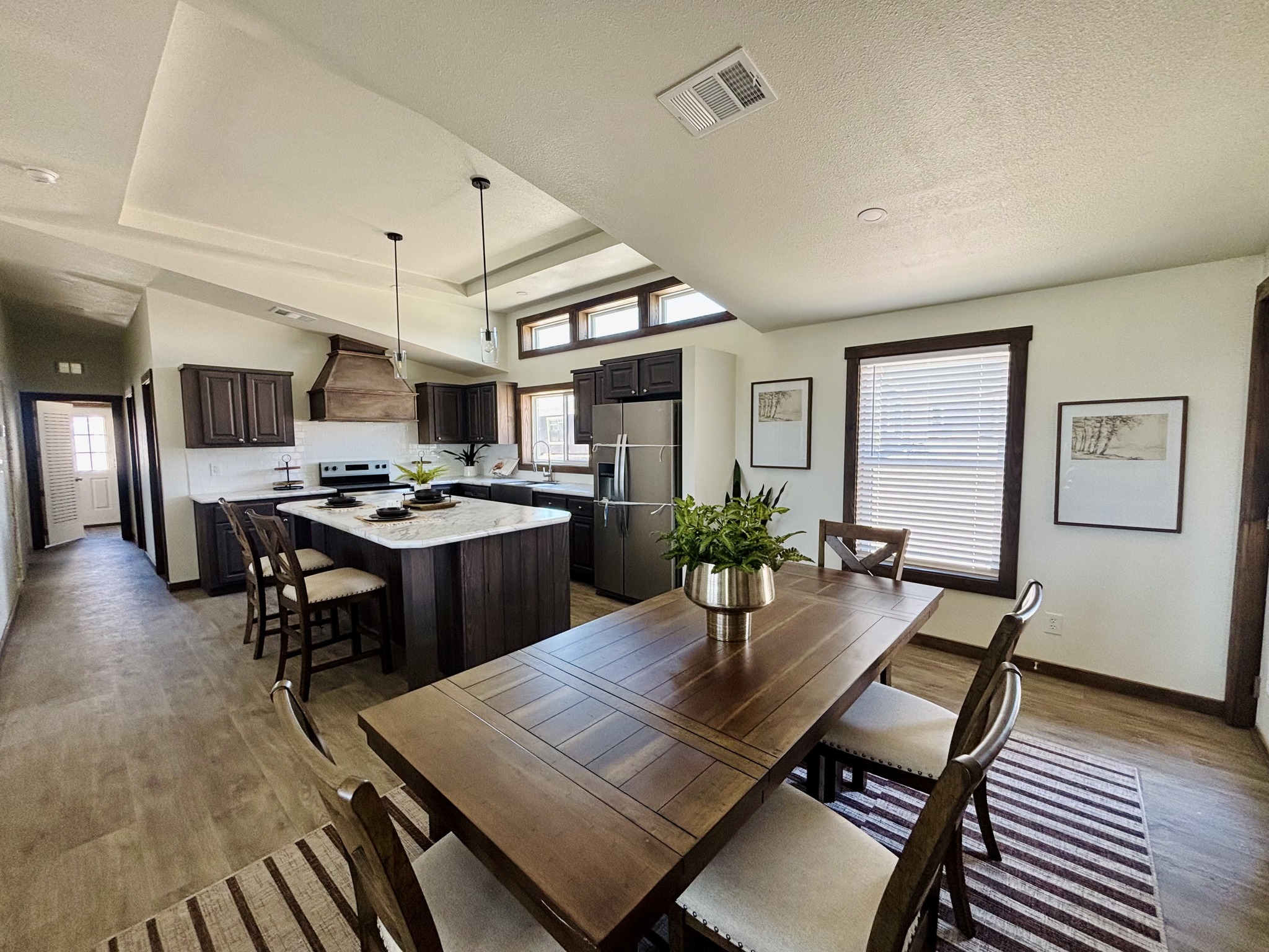 Modern kitchen and dining area with warm tones. Features include a wooden dining table, chairs, island with barstools, stainless steel appliances, and large windows for natural light.